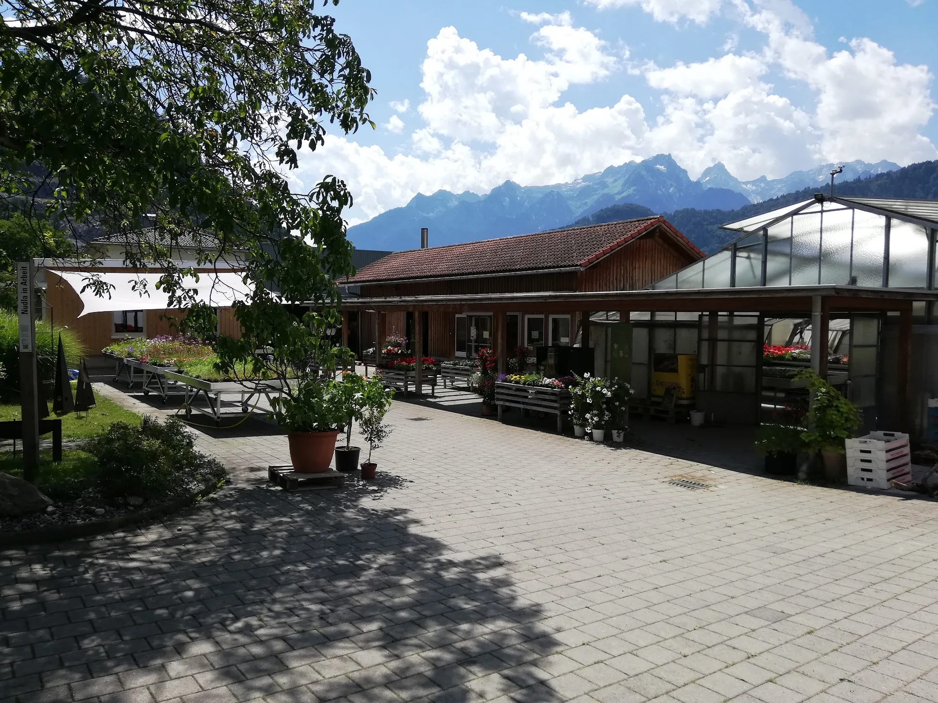 Der Hof der Gärtnerei Hartmann mit Verkaufsgebäude, Glashaus und Blumentischen vor der Bergkulisse